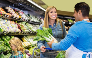 Woman shopping in a grocery store with her young child. She is talking to a grocery store employee who is helping her select lettuce.